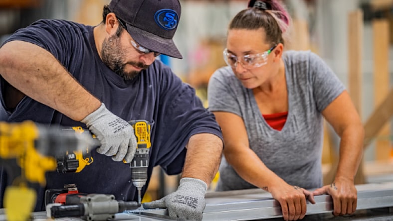 Two Pallet employees, a man and a woman, working on the Pallet production line. The man (left) is wearing a navy blue t-shirt and work gloves, using a power drill to drill a hole in a shelter component as the woman (right)—wearing a grey t-shirt and holding the other end of the component being worked on—looks on. Both individuals are wearing safety glasses.