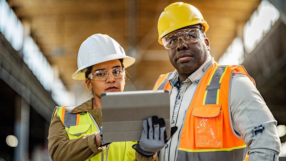 A man and a woman in safety gear looking at tablet on construction site.