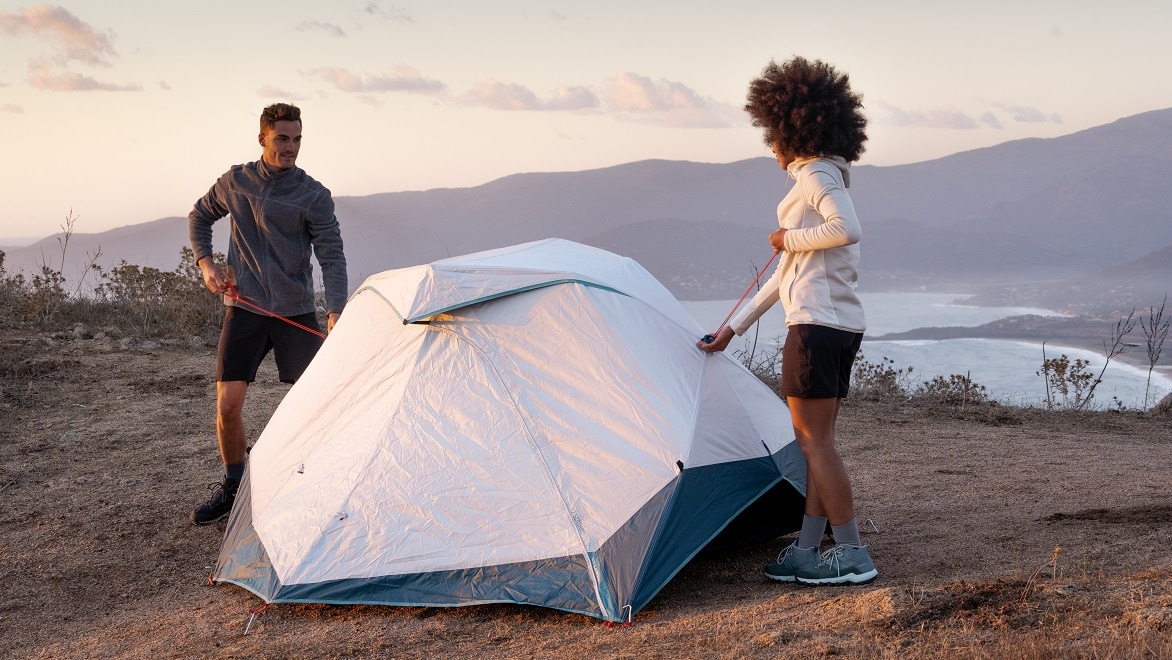 Zwei Personen bauen ein Campingzelt über einem Abhang und mit Blick auf einen See auf.