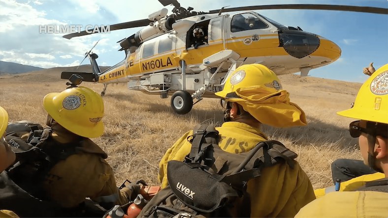 A view from the helmet cam of a firefighter shows several firefighters in full gear in front of an L.A. Country Fire Department helicopter in a field.