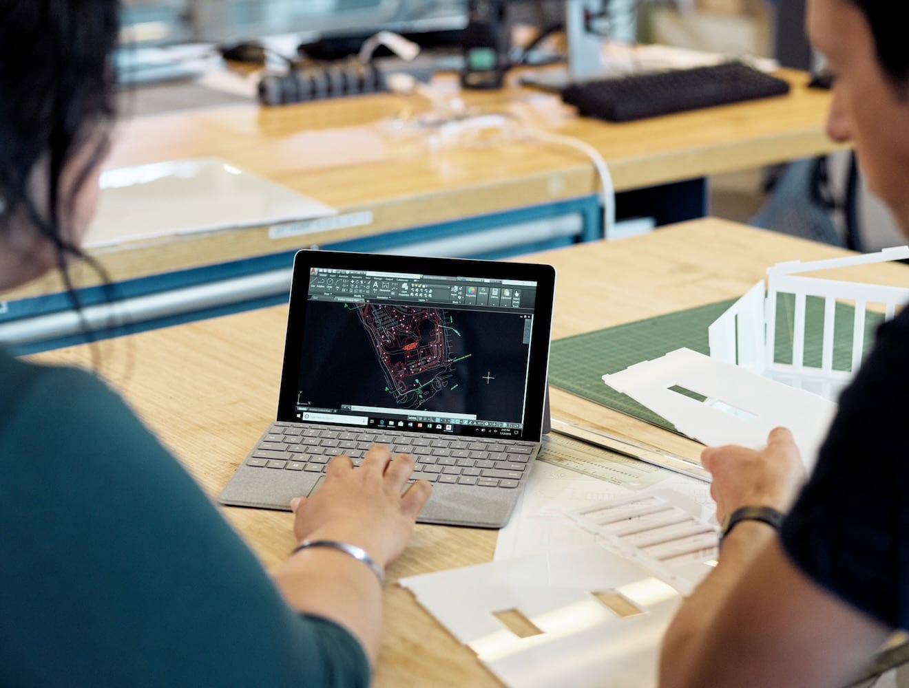 A view over the shoulders shows two people working on a tablet with an attached keyboard displaying AutoCAD.