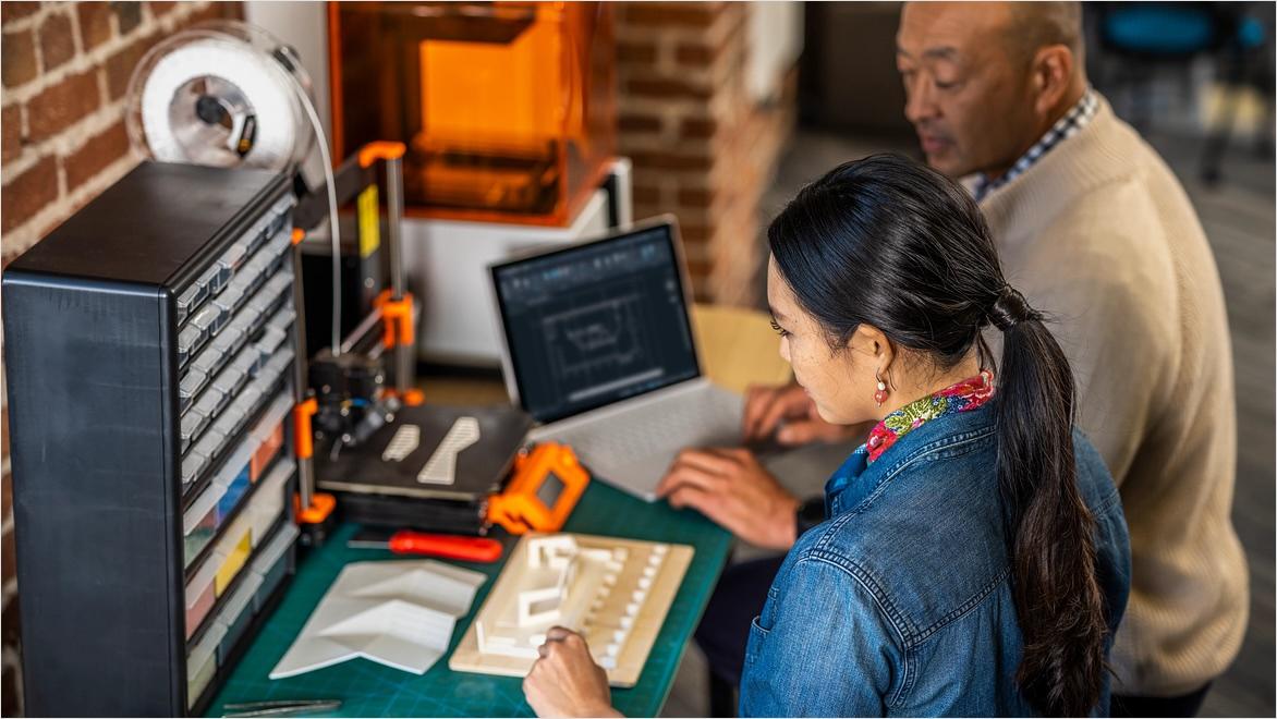 A man and a woman in a maker space look at a 3D-printed model alongside a laptop displaying its design in AutoCAD.