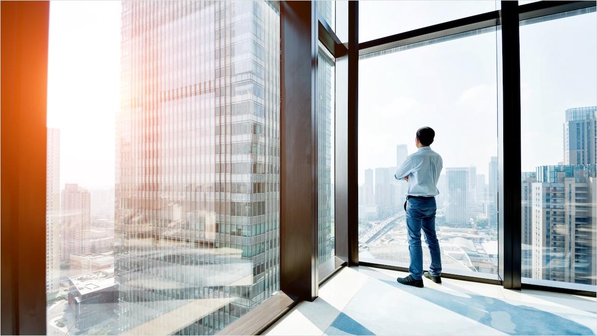 A businessman stands at an expansive window looking out at a skyscraper.