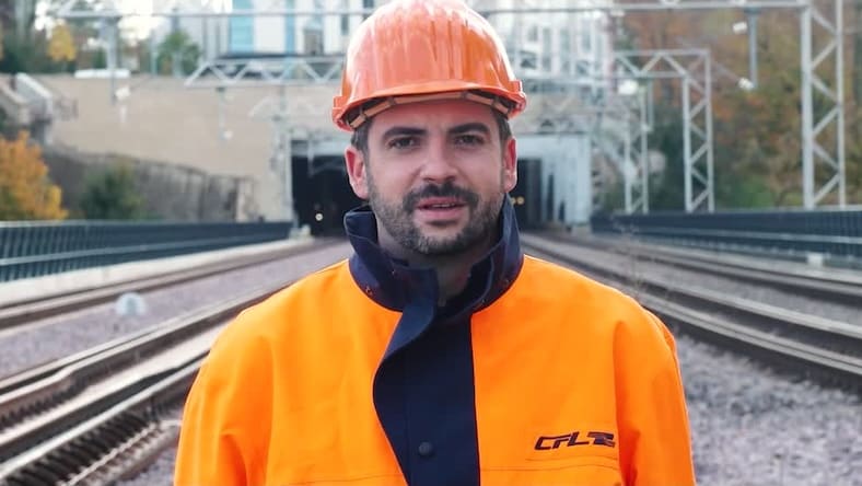 A man in an orange hard hat and jacket stands on train tracks.