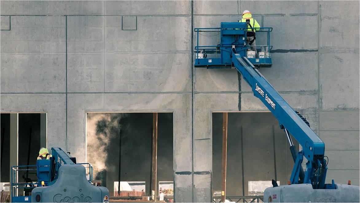 Two construction workers on elevated platforms in front of concrete wall