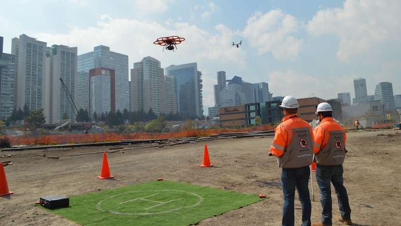 Two men on a construction site operate flying drones.