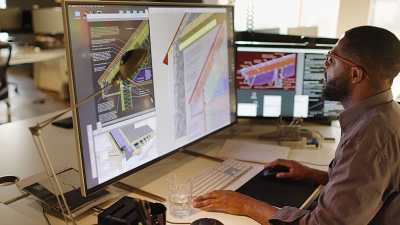 A man examines detailed architectural and engineering designs displayed on a large computer monitor in an office setting.