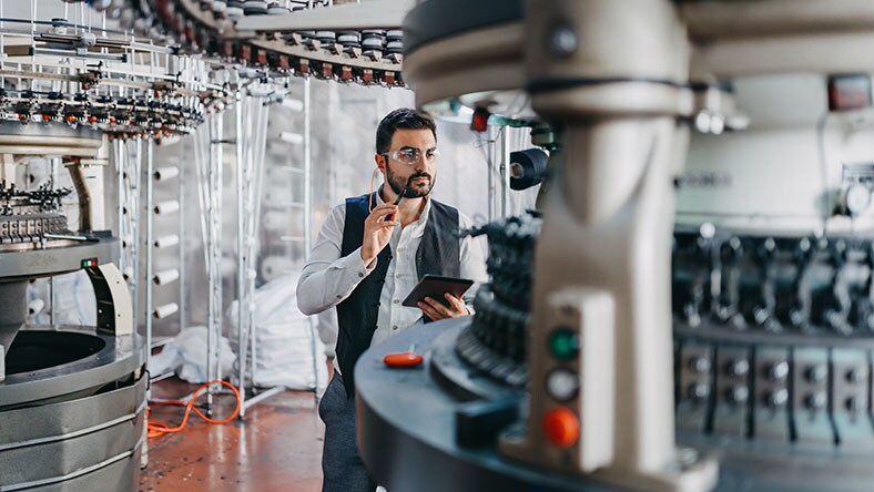 A man uses a tablet to inspect industrial textile machinery in a manufacturing facility.