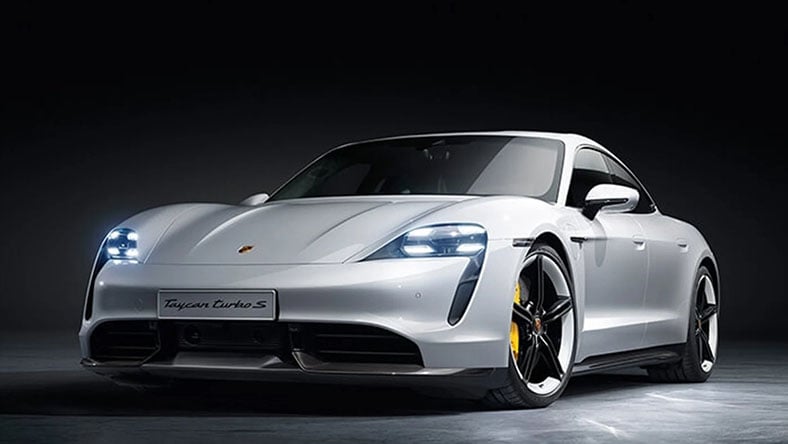 A white Porsche stands in a dark studio with its headlights illuminated.