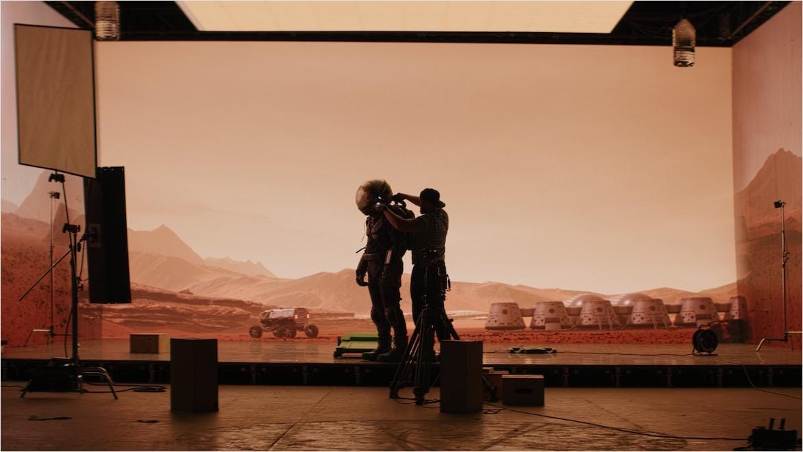 An actor dressed as an astronaut stands on a TV set as a member of the production team adjusts the suit.