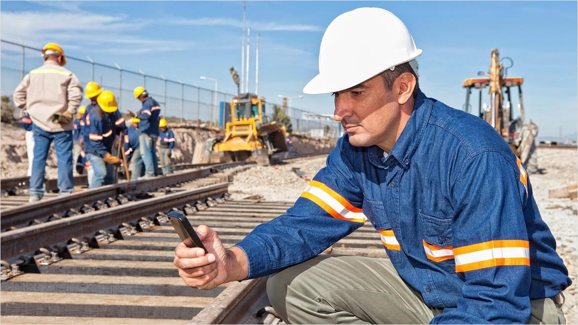 An engineer wearing a hard hat kneels to use digital technology next to a rail at a construction site.