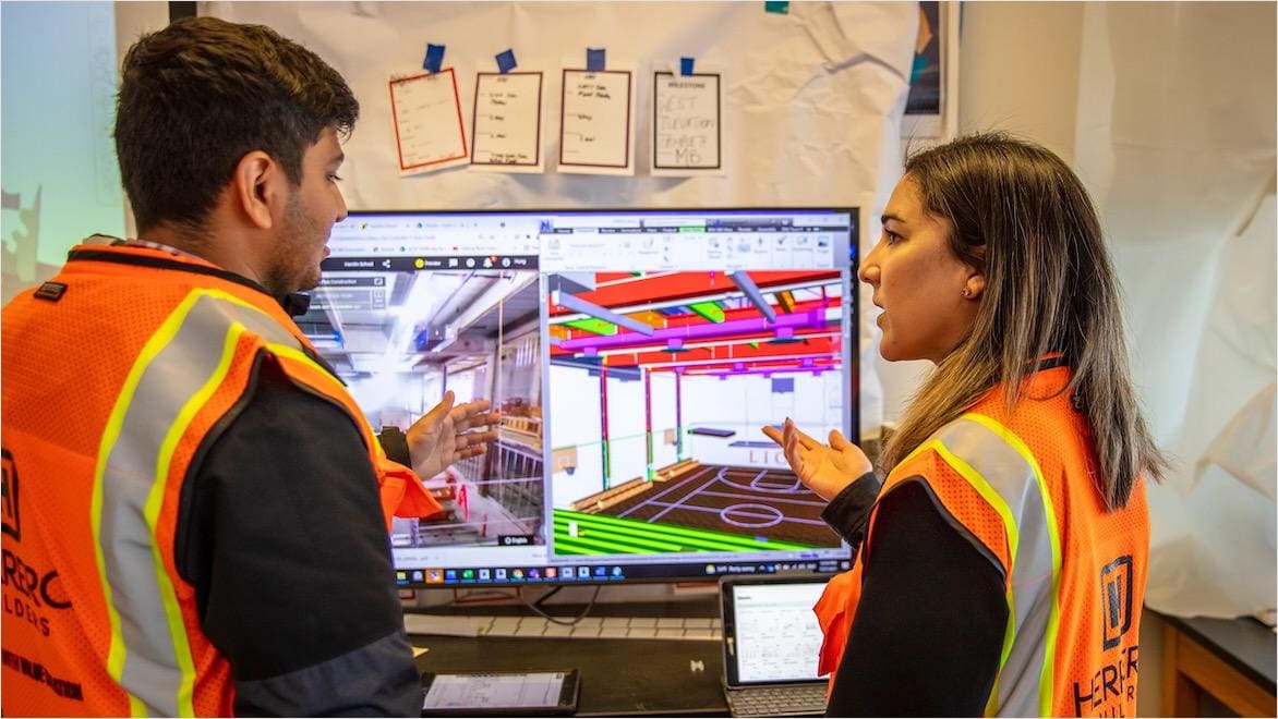 A man and woman in safety vests consult a computer screen displaying a building model.