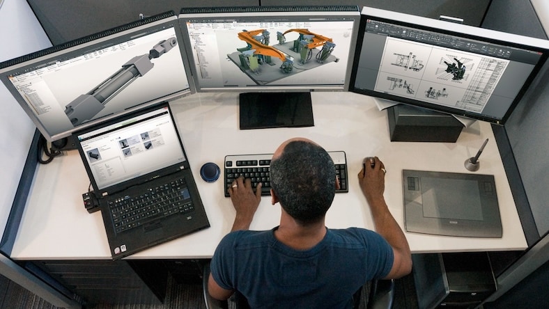A man sits at a desk with multiple digital devices, including a computer, screens, and mobile devices.