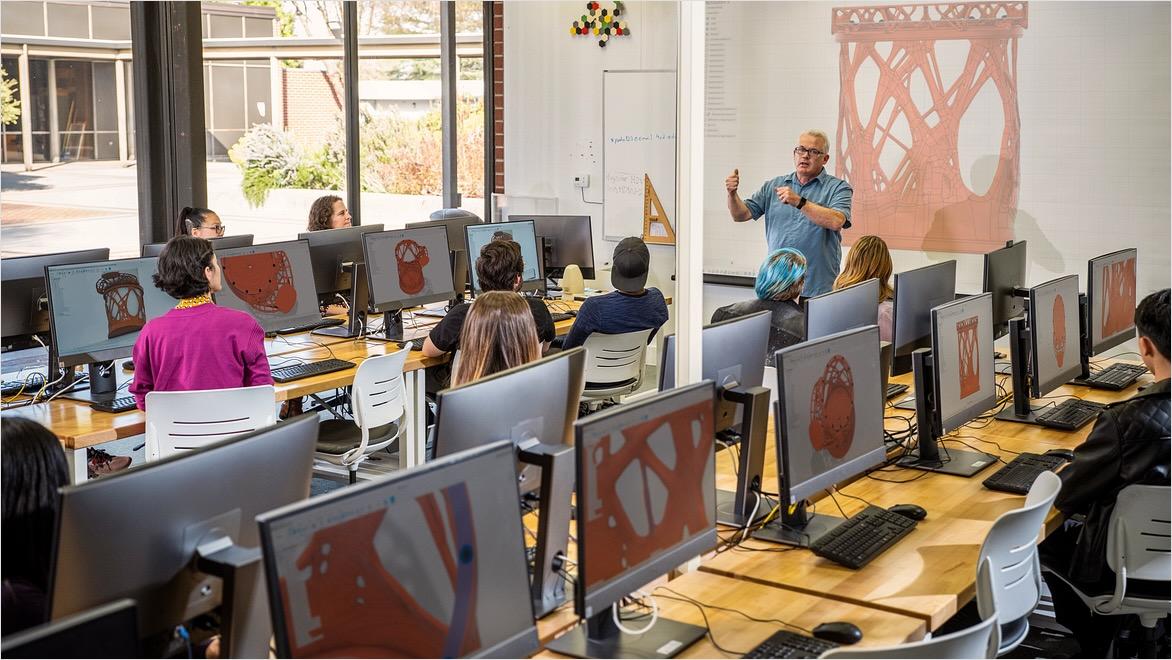 A college instructor stands in front of a screen displaying a design in Fusion software; the students sit in front of computer monitors displaying the same design.