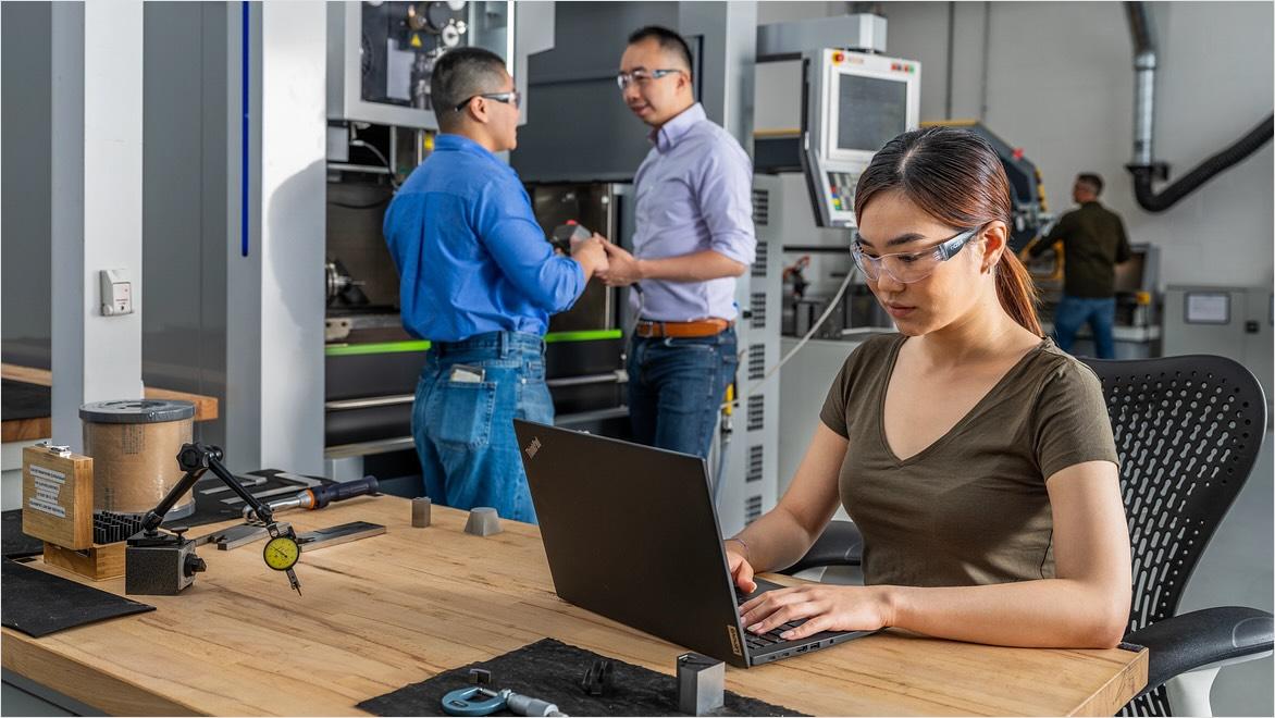 A woman wearing safety glasses works on a laptop in a machine shop while other people stand by machines talking.