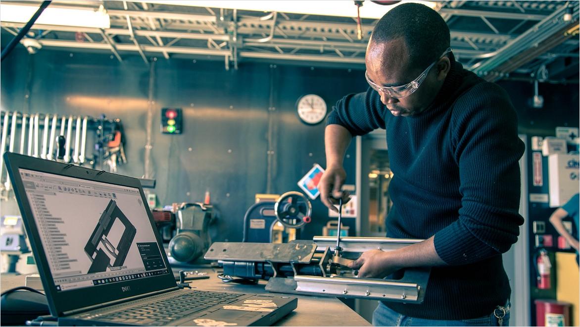 A man works in a machine shop next to a laptop displaying Fusion software.