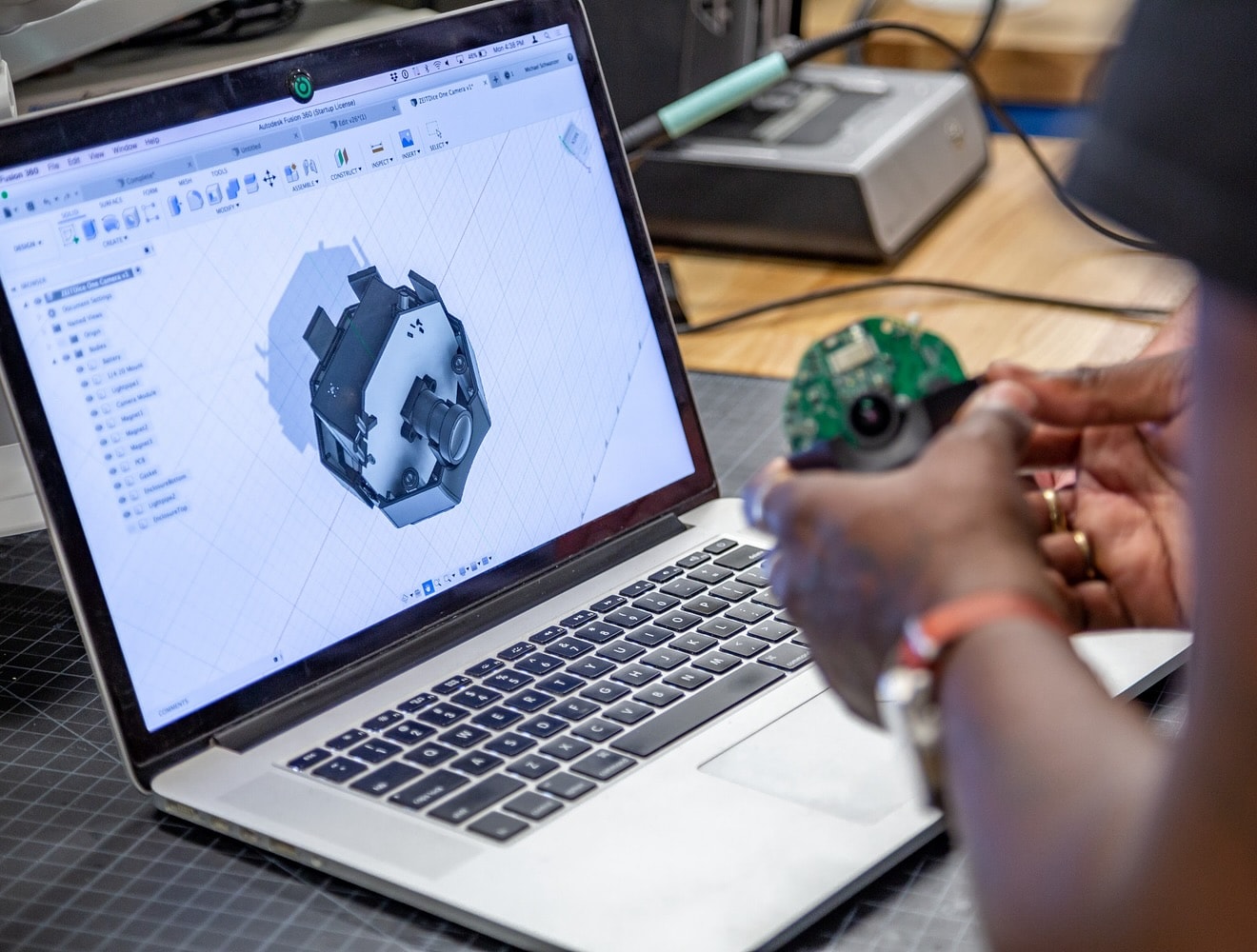 A close-up of a man’s hands shows him holding a small device in front of a laptop computer displaying the device’s design in Fusion software.