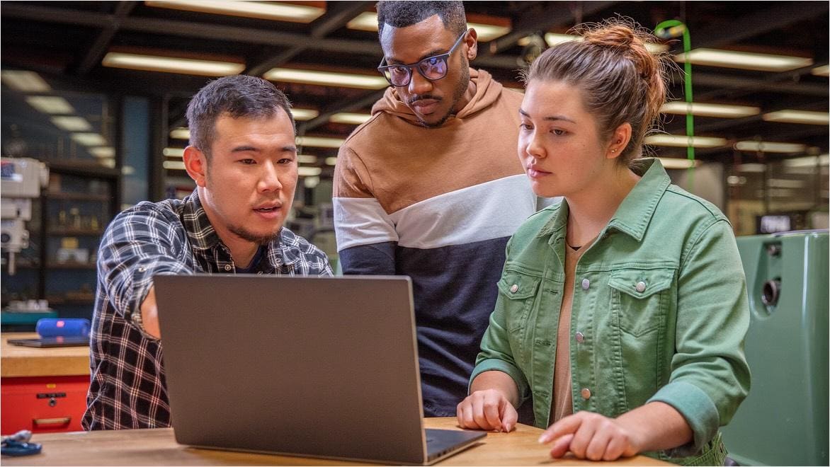 A teacher and two young adult students in a CNC machining class look at a laptop.
