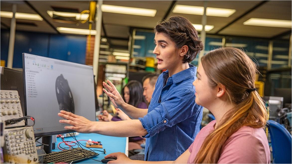 In a CNC electronics lab, an instructor works with a student at a computer displaying Fusion software.