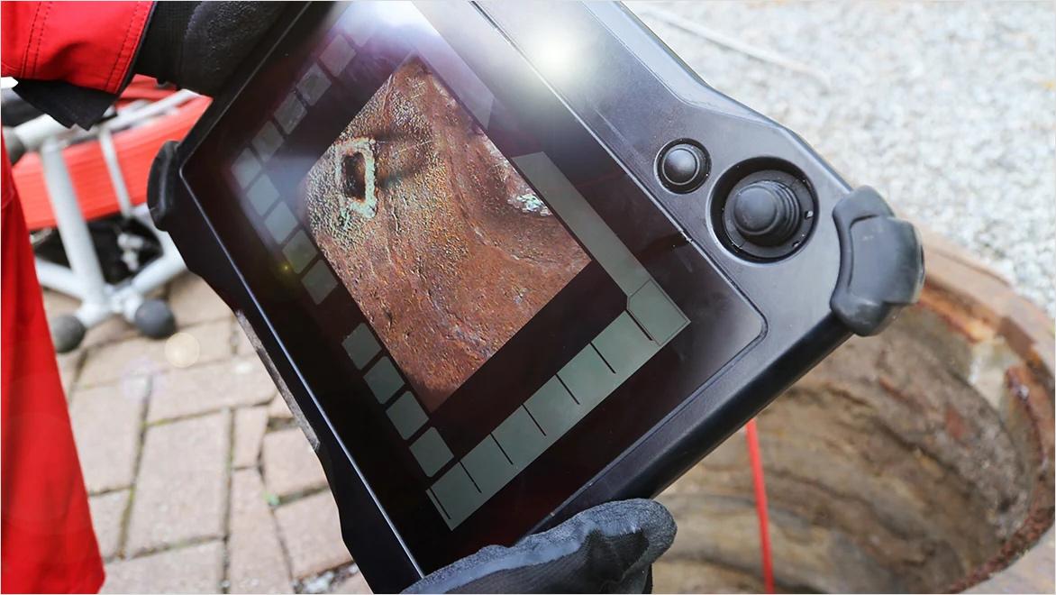 A handheld tablet shows footage from a camera inside an open drainpipe.