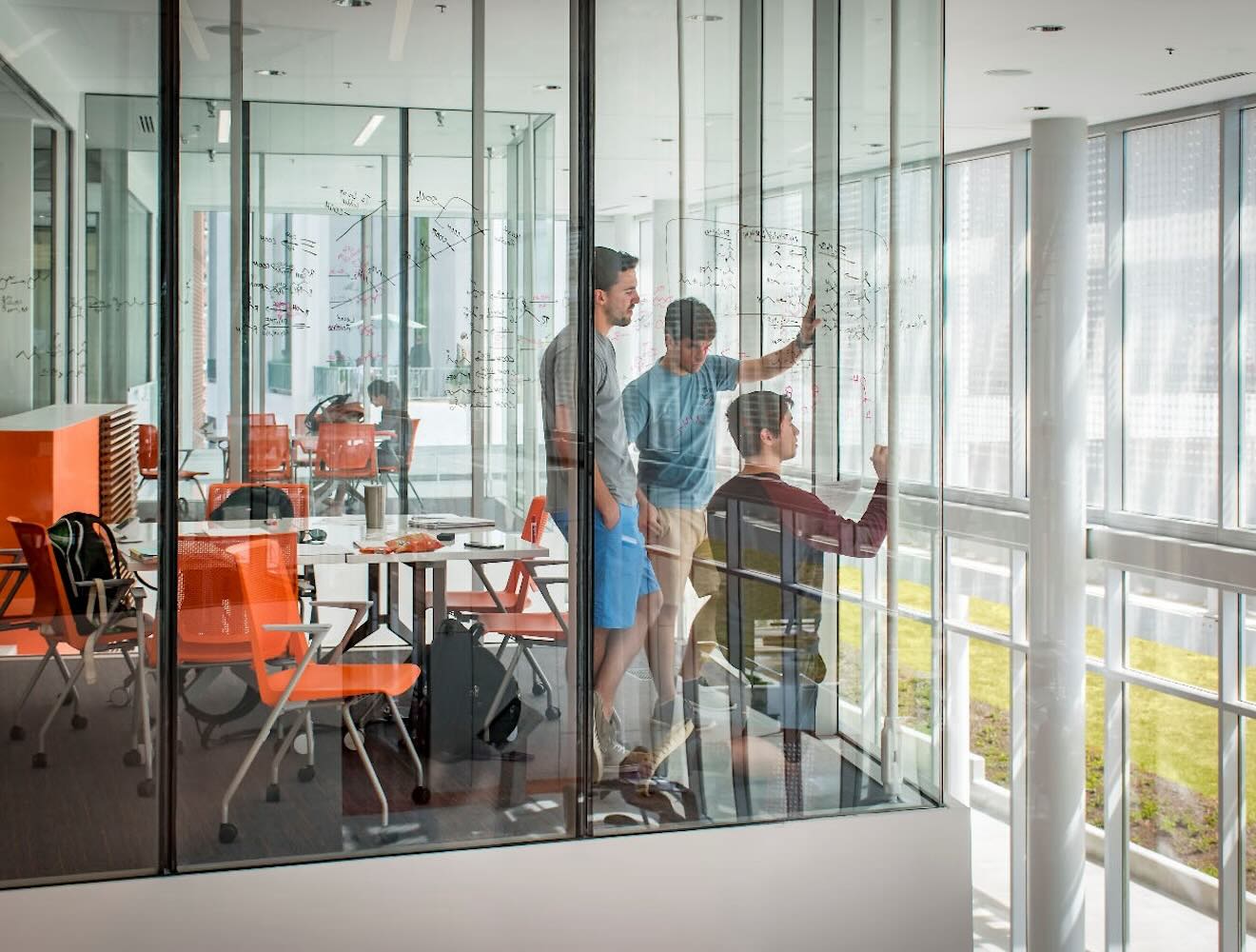 Three people stand inside a glass-walled room, writing equations on the glass, with orange chairs and desks visible in the background.