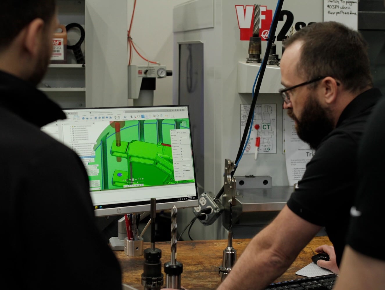 Two men in a machining shop look at a computer monitor displaying a screen from Autodesk Fusion.