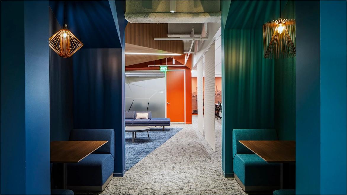 A view of a modern office shows conversation booths with warm lighting on either side of a hall leading to a brightly lit common area.