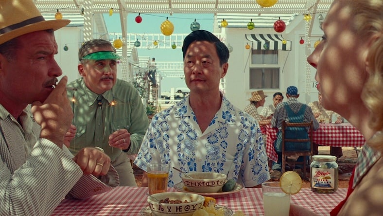 A still from the film Asteroid City shows several people in 1950’s attire sitting at a picnic table under a white pergola strung with colorful lights.