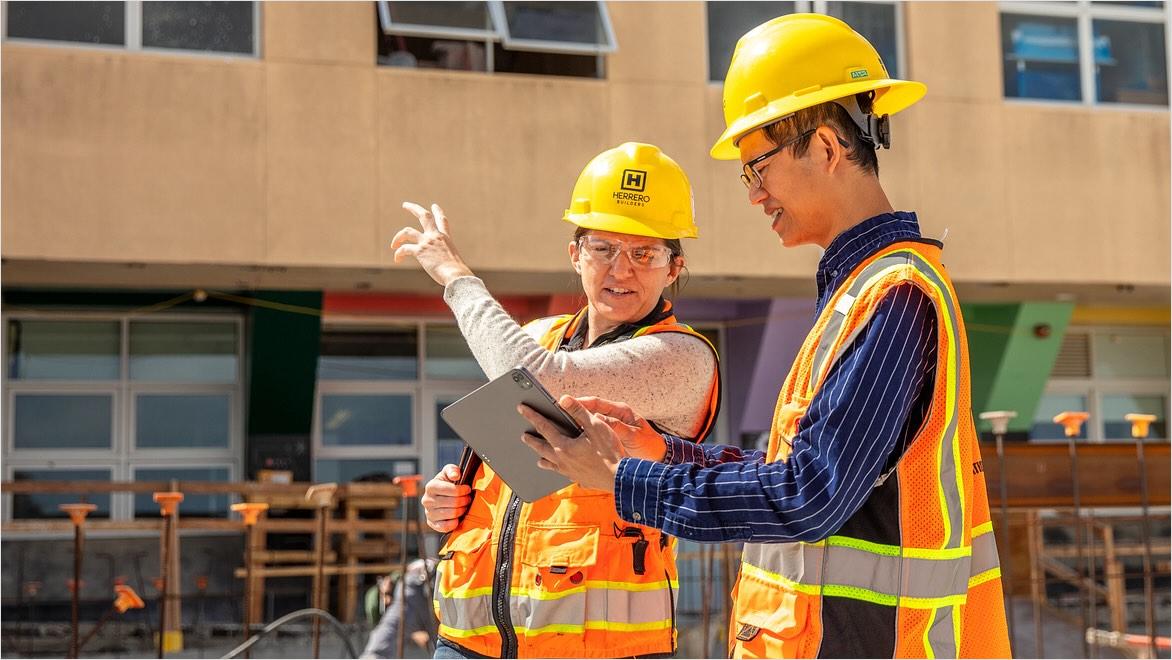 Two people wearing safety gear on a construction site look at a tablet.