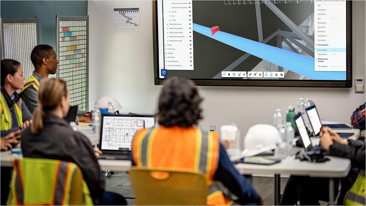 A group of construction workers in safety vests consult a building model on a large monitor.