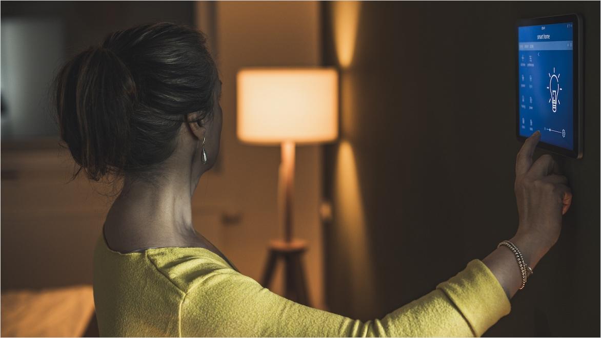 A woman uses a wall-mounted tablet to adjust lighting in a home.