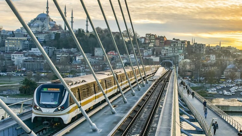 A modern train travels on a bridge over water with a cityscape of Istanbul in the background.