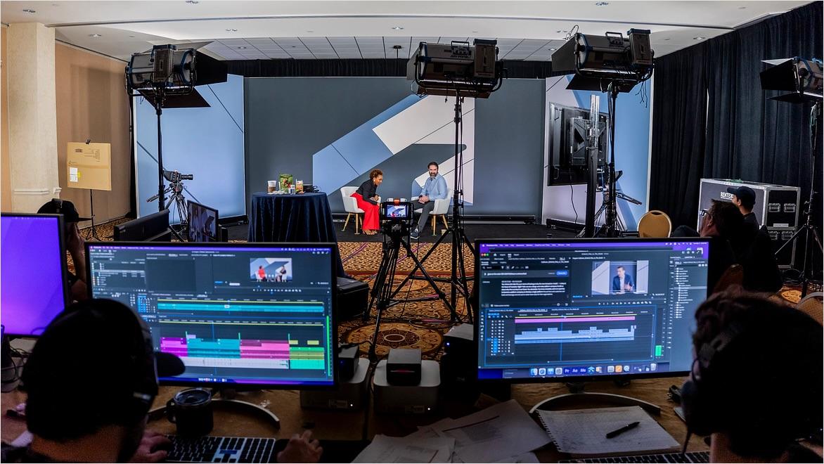 A recording set up at a conference includes lighting, cameras, microphones, and computer monitors for video and audio production.