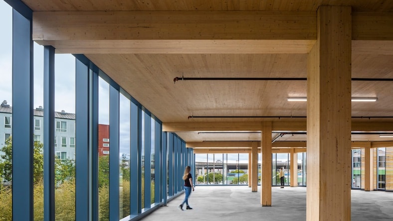 A vast building interior is defined by large wood beams and floor-to-ceiling windows.