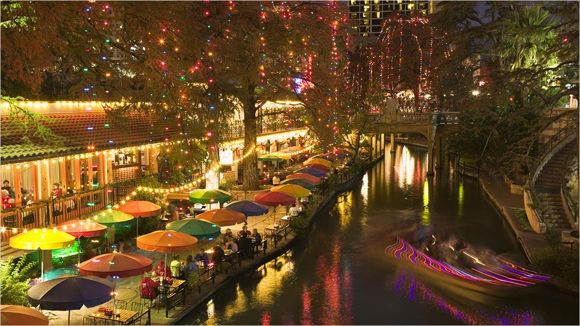 An urban restaurant-lined riverfront is shown at night.