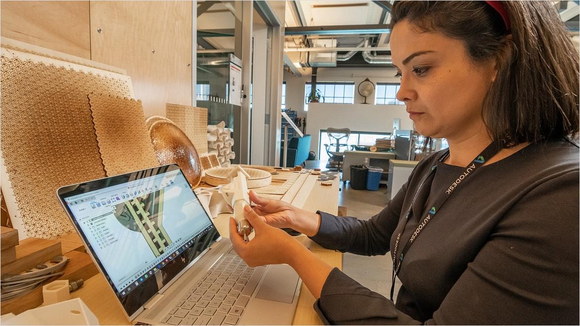 A woman holds a 3D-printed object while looking at a laptop displaying the CAD for the object in Fusion.