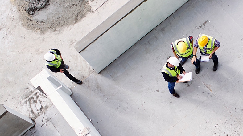 Workers consult plans while standing on a concrete foundation. 
