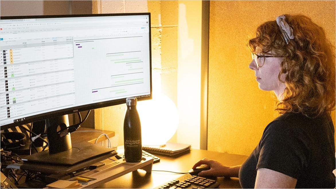 A woman sits at a computer screen displaying a timeline of media assets.