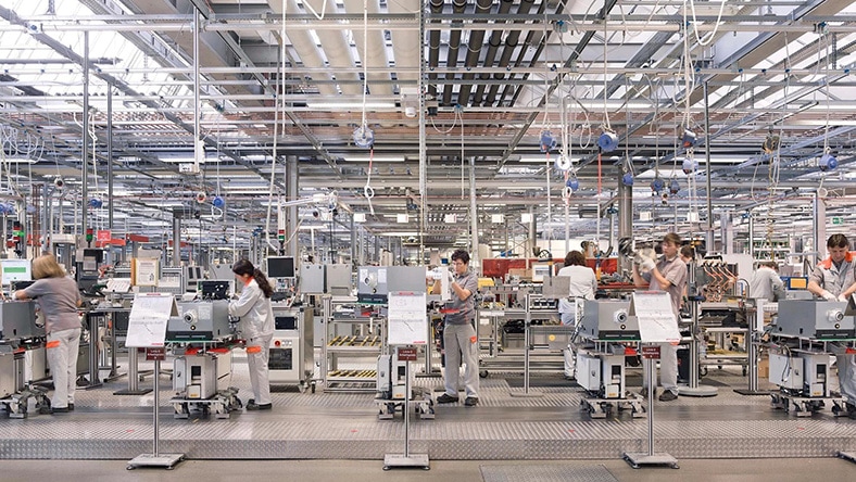 Workers stand at automated stations in a factory.