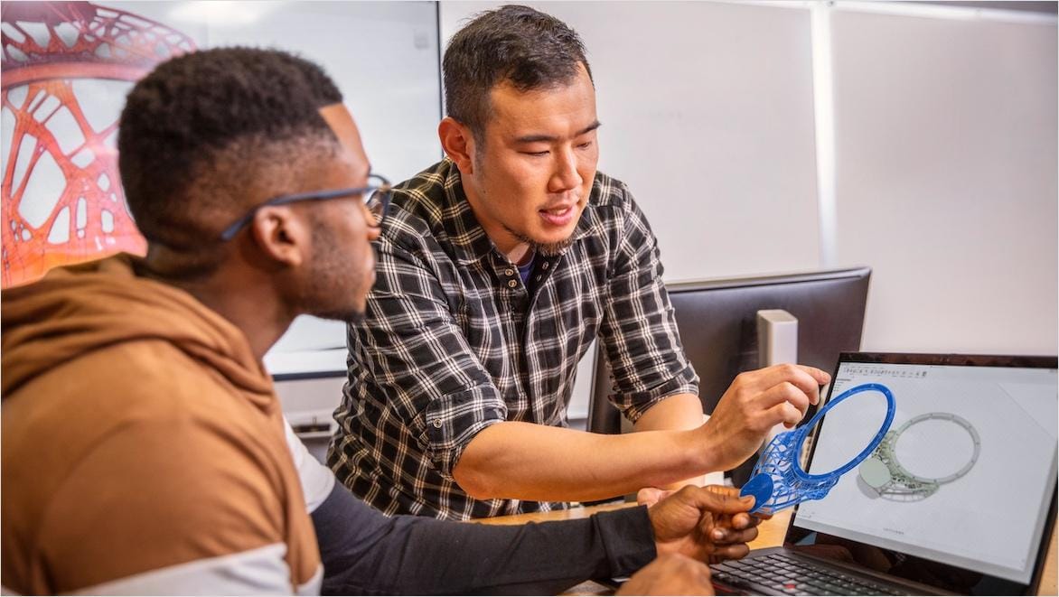 Two men compare an intricate 3D printed part to the design on a computer screen.