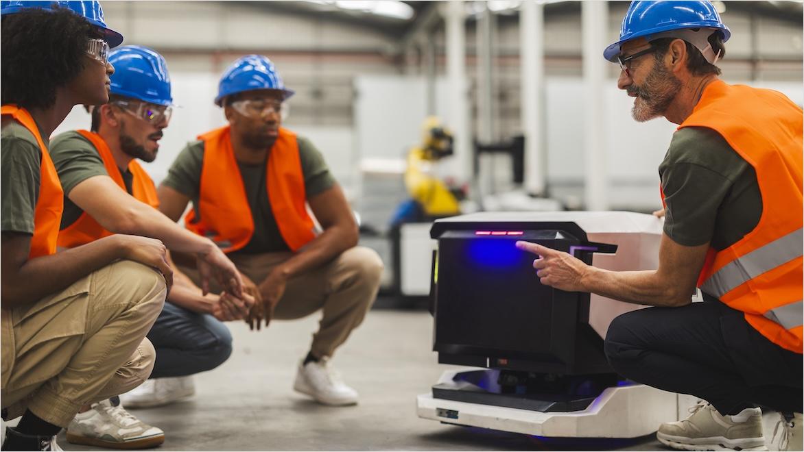 Three people in safety vests and hard hats crouch on the floor of a factory as another man shows them how to operate an automated guided vehicle.