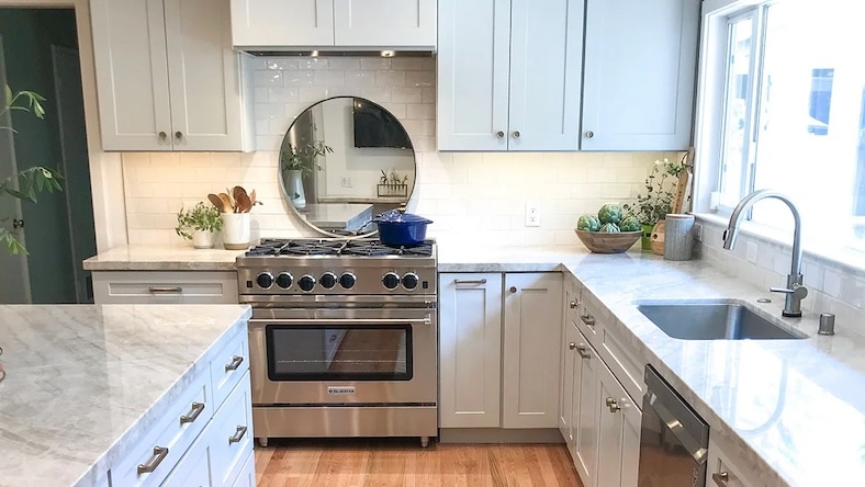 A redesigned kitchen includes a mirror behind the range, light marble counters, and hardwood floors.