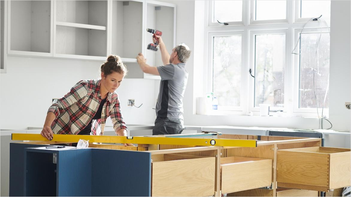 A woman uses a level on kitchen island cabinets prior to installing a countertop. Her male colleague uses a cordless drill on wall cabinet frames.
