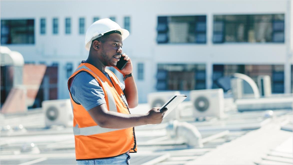 A man in a hard hat standing on a building roof consults data on an iPad