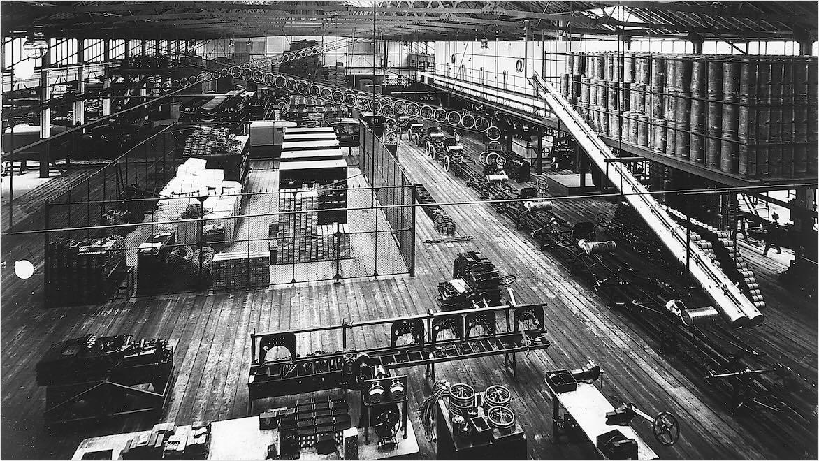 A black and white photo shows Ford Model T automobiles being built on an assembly line, circa 1914.