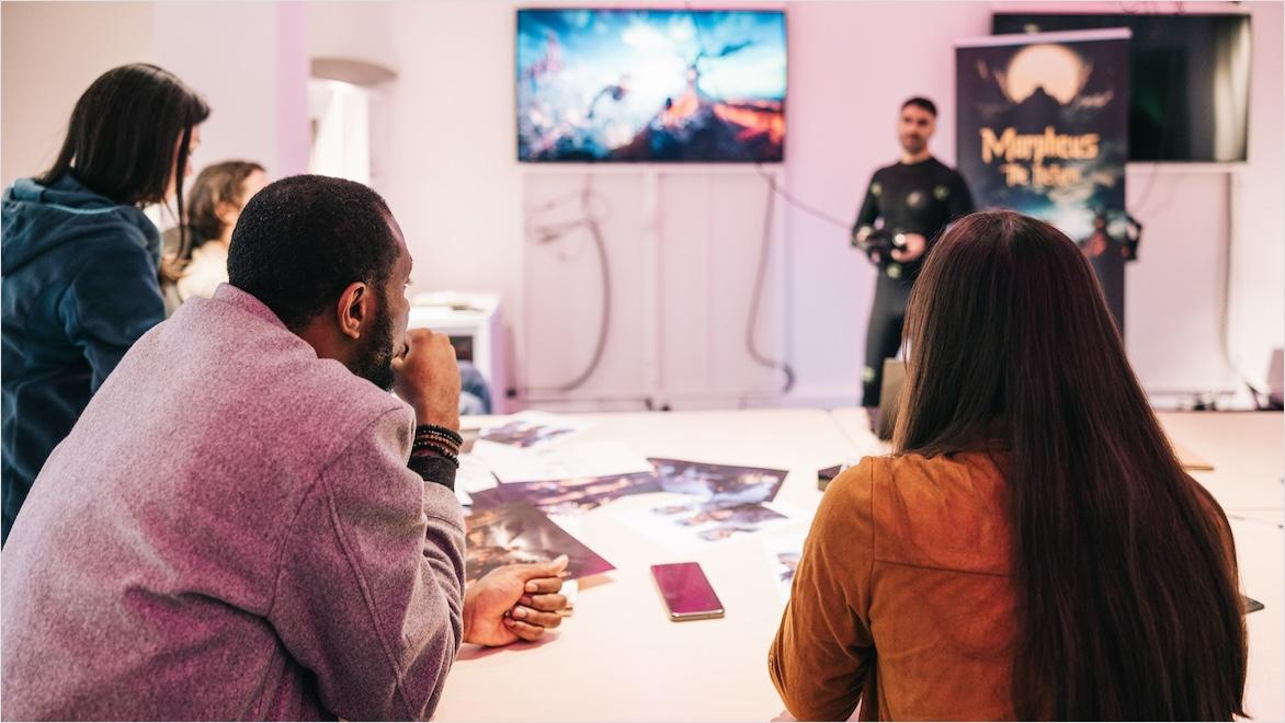A group of game designers sit at a conference table and view action on a screen.