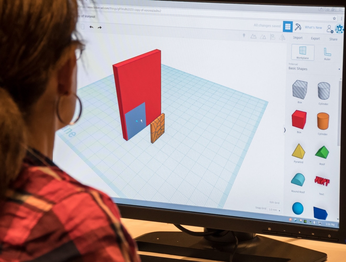 A view over the shoulder of a woman shows her using TinkerCAD on a desktop computer.