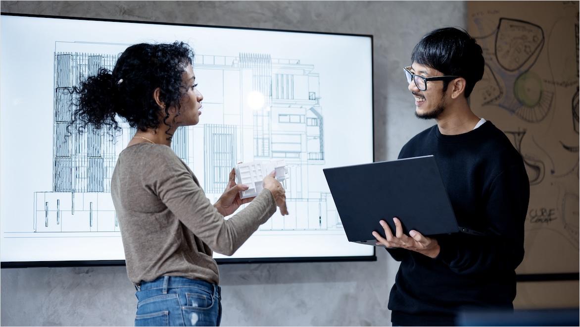 A woman and man discuss a construction project in front of a large screen showing a building blueprint.