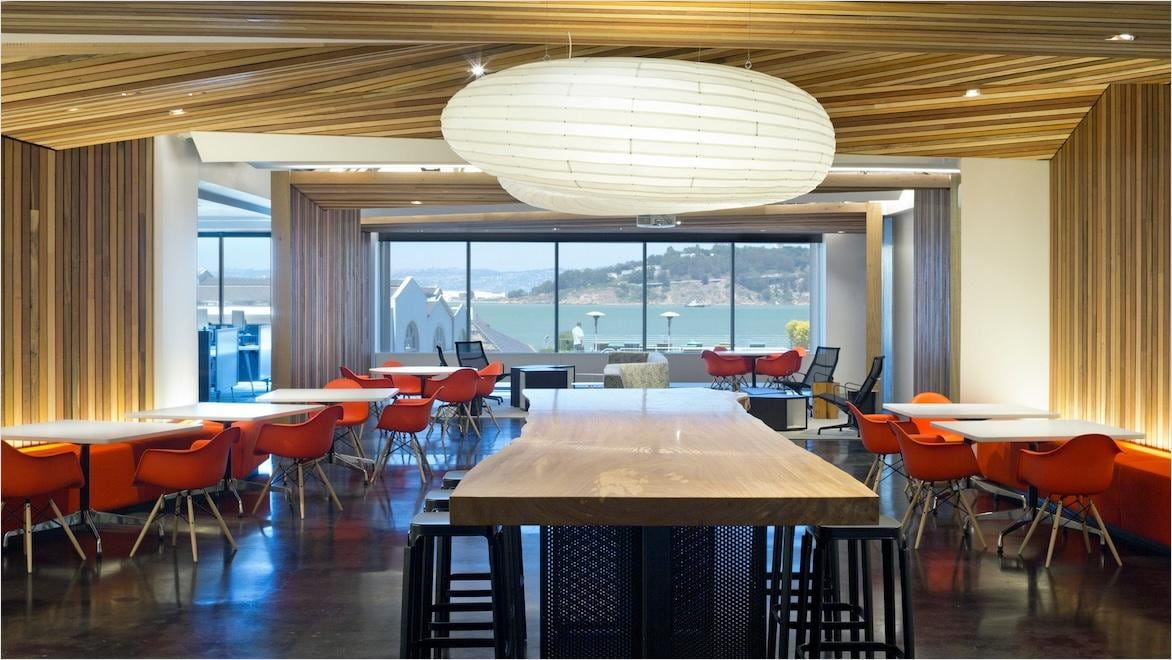 A dining area in a modern office has a large modern light fixture over a central table, downlights, and uplights throughout to produce ambient light and highlight architectural features.