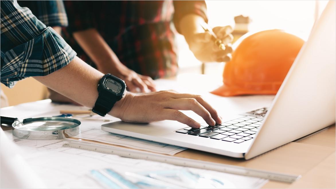 Two architects use a laptop; hard hats sit on the table.